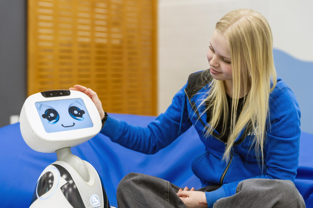 Nuori nainen istuu ja hymyilee katsellen pientä robottia, jonka näytöllä on iloiset kasvot.