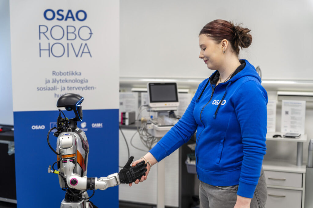 Nainen sinisessä hupparissa kättelee robottia. Taustalla on OSAO ROBOHOIVA -kyltti ja terveydenhuollon laitteita.