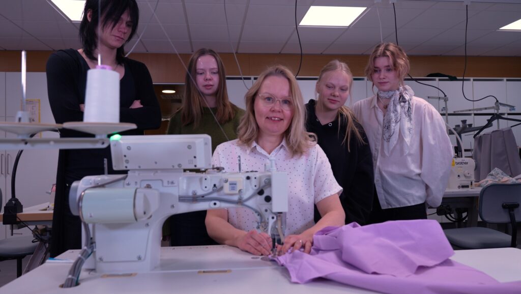 Kuvassa nainen istuu ompelukoneen ääressä hymyillen, kun neljä nuorta henkilöä seisoo hänen takanaan katsellen. He ovat sisätiloissa, ja pöydällä on violettia kangasta.