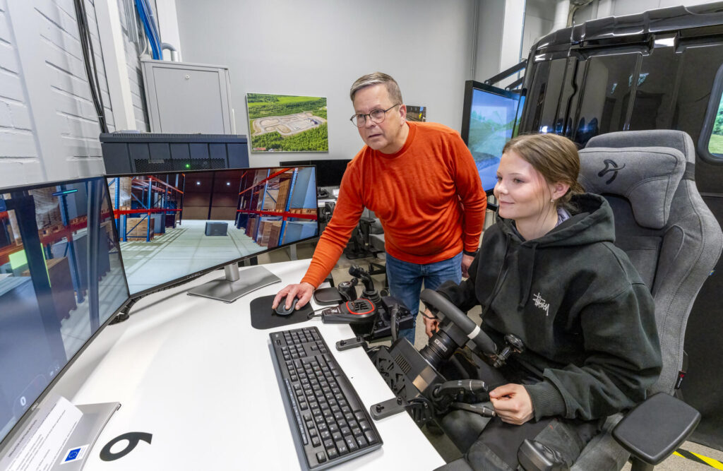 Kaksi henkilöä on ajosimulaattorin ääressä. Opiskelija käyttää simulaattorilaitteistoa, joka on yhdistetty suuriin näyttöihin, ja opettaja neuvoo häntä. Näytöillä näkyy virtuaalinen ympäristö, joka muistuttaa varastoa tai tehdasta. Huoneessa on myös muita näyttöjä ja laitteita.
