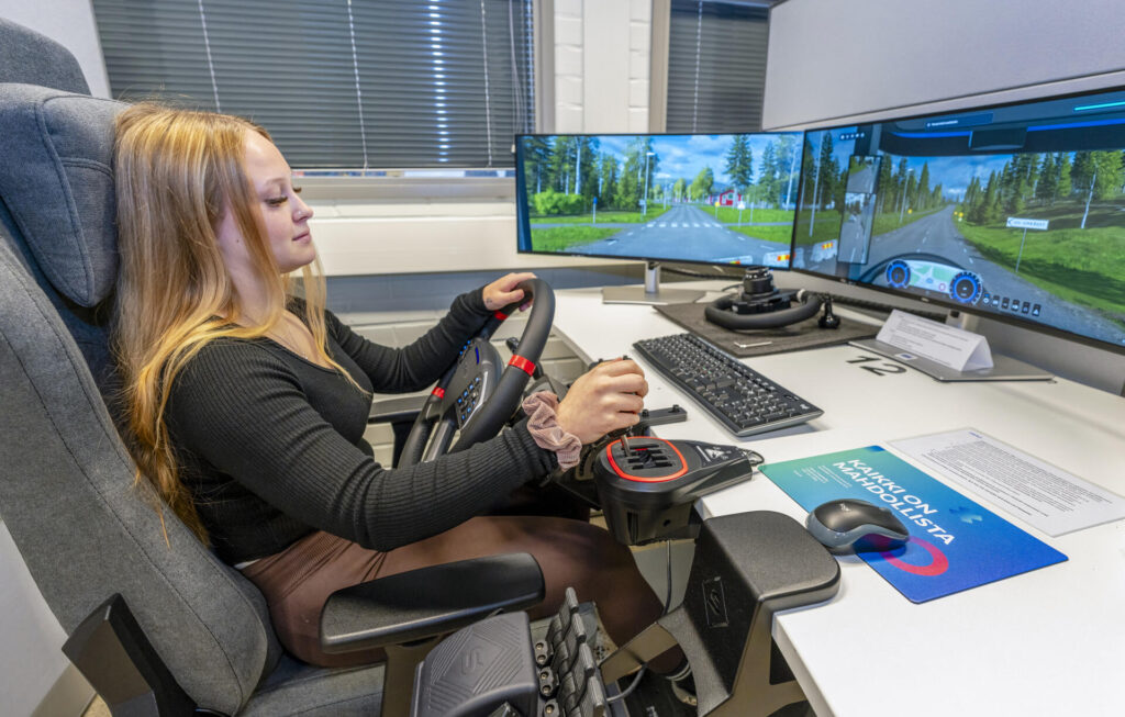 Opiskelija käyttää ajosimulaattoria toimistoympäristössä, jossa pöydällä on kaksi tietokoneen näyttöä, jotka näyttävät virtuaalisen tieympäristön.