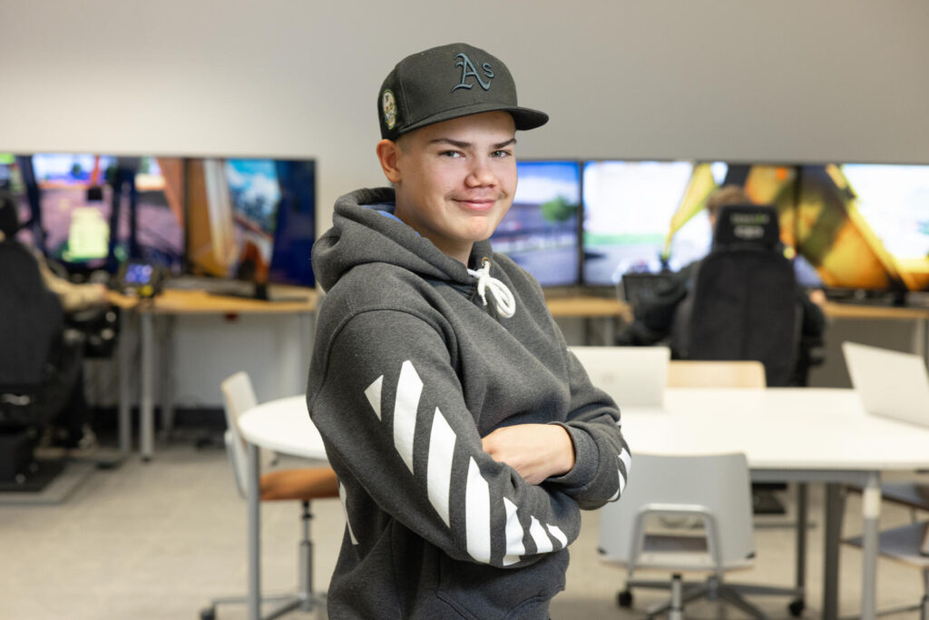 Henkilö seisoo luokkahuoneessa, jossa on useita näyttöjä taustalla. Hänellä on harmaa huppari ja lippalakki. Ympärillä on pöytiä ja tuoleja, ja yksi toinen henkilö istuu tietokoneen ääressä.