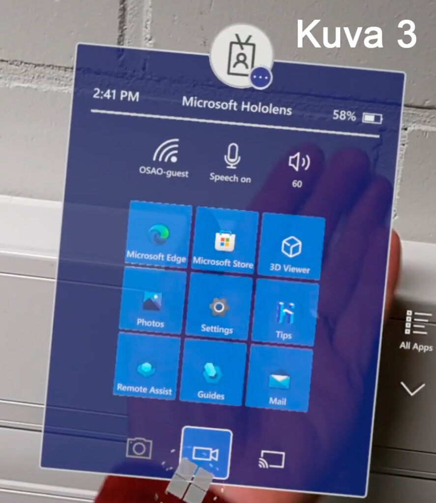 Microsoft Hololens -käyttöliittymä, jossa näkyy sovelluskuvakkeita kuten Microsoft Edge, Microsoft Store, 3D Viewer, Photos, Settings, Tips, Remote Assist, Guides ja Mail. Näytöllä on myös Wi-Fi-, puhe- ja äänenvoimakkuusikonit. Kuvan otsikko on 'Kuva 3'.