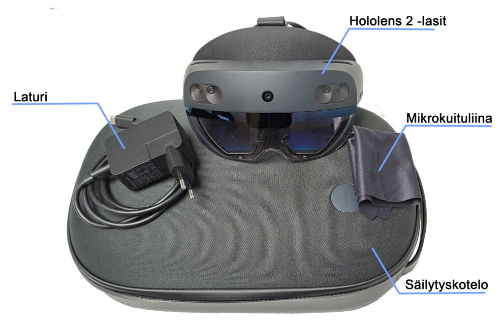 Kuvassa on HoloLens 2 -lasit, jotka on sijoitettu mustan säilytyskotelon päälle. Kuvassa näkyy myös laturi, joka on sijoitettu vasemmalle puolelle, sekä mikrokuituliina oikealla puolella.