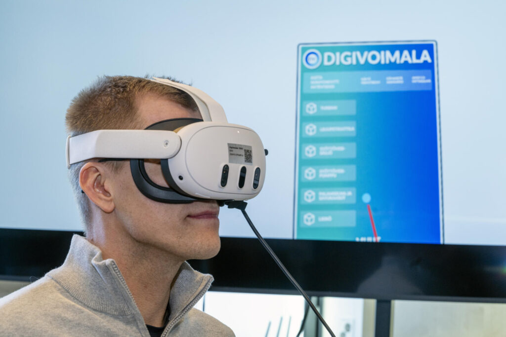 Henkilö käyttää virtuaalitodellisuuslaseja, taustalla näkyy näyttö, jossa lukee 'DigiVoimala'.