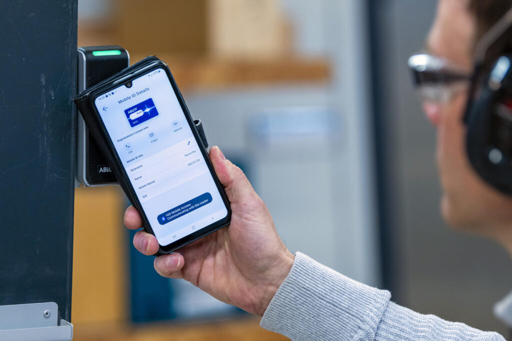 Henkilö käyttää älypuhelinta digitaaliseen tunnistautumiseen oven lukijalaitteen avulla. Puhelimen näytöllä näkyy sovellus, jossa on sähköinen tunnistautuminen ja valikkoja.