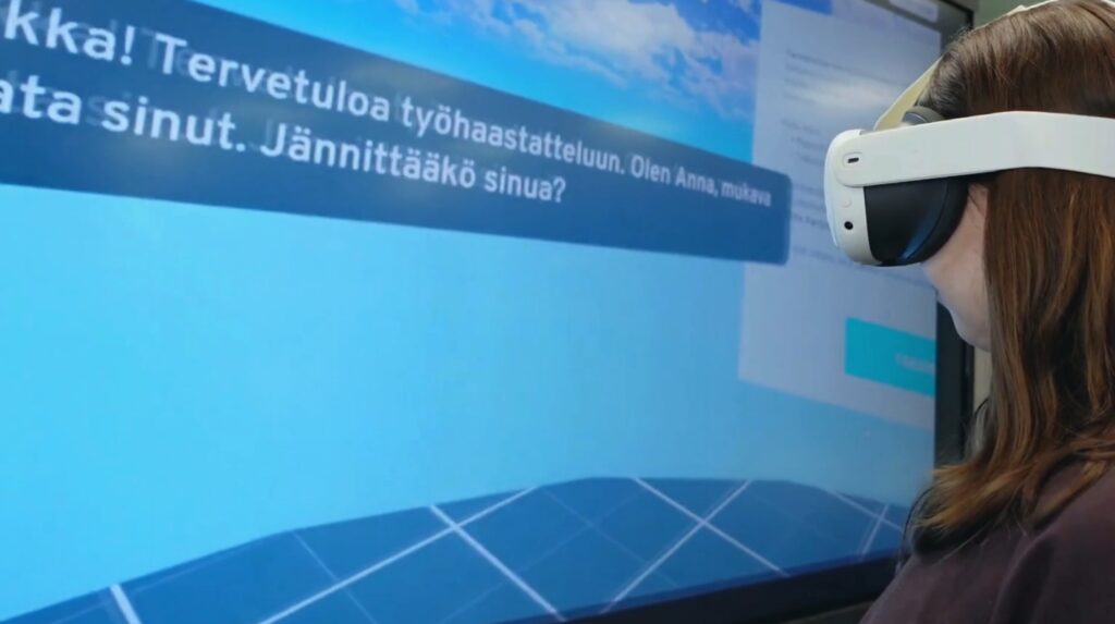 Henkilö käyttää virtuaalitodellisuuslaseja ja katsoo näyttöä, jolla lukee: 'Terve! Tervetuloa työhaastatteluun. Olen Anna, mukava tavata sinut. Jännittääkö sinua?' Näytöllä on sininen tausta ja pilviä yläosassa.