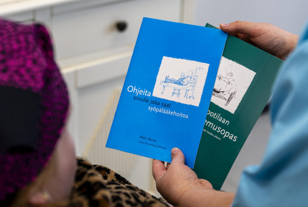 Kaksi henkilöä pitelee kahta kirjaa käsissään. Ensimmäinen kirja on sininen ja sen kannessa lukee "Ohjeita sinulle joka saat syöpälääkehoitoa". Toinen kirja on vihreä ja sen kannen otsikosta on näkyvissä vain osa, sekä piirroskuva, joiden perusteella voi päätellä kirjan nimen olevan "Syöpäpotilaan ravitsemusopas".