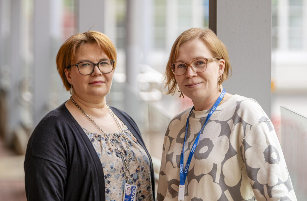 Kaksi henkilöä seisoo rinnakkain sisätiloissa. Molemmilla on silmälasit ja vaaleat hiukset. Heillä on yllään tyylikkäät vaatteet ja kaulassa roikkuu sininen OSAOn nimikyltti.