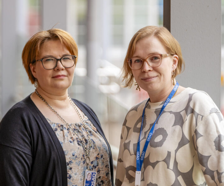 Kaksi henkilöä seisoo rinnakkain sisätiloissa. Molemmilla on silmälasit ja vaaleat hiukset. Heillä on yllään tyylikkäät vaatteet ja kaulassa roikkuu sininen OSAOn nimikyltti.