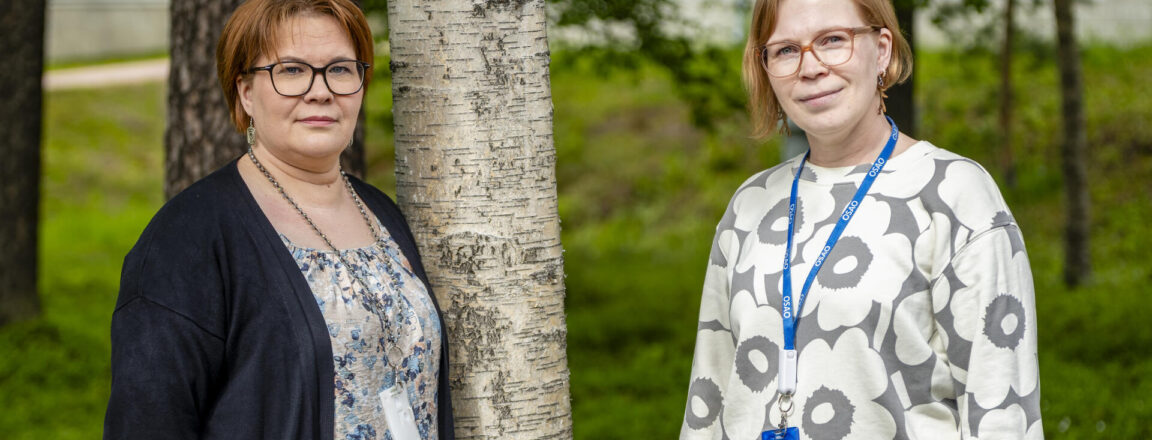 Kaksi henkilöä seisoo ulkona vierekkäin, taustalla on puita ja vehreää ympäristöä. Heillä on yllään rento vaatetus ja kaulanauhat, joissa roikkuu kulkukortit.