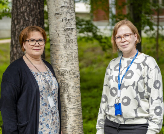 Kaksi henkilöä seisoo ulkona vierekkäin, taustalla on puita ja vehreää ympäristöä. Heillä on yllään rento vaatetus ja kaulanauhat, joissa roikkuu kulkukortit.