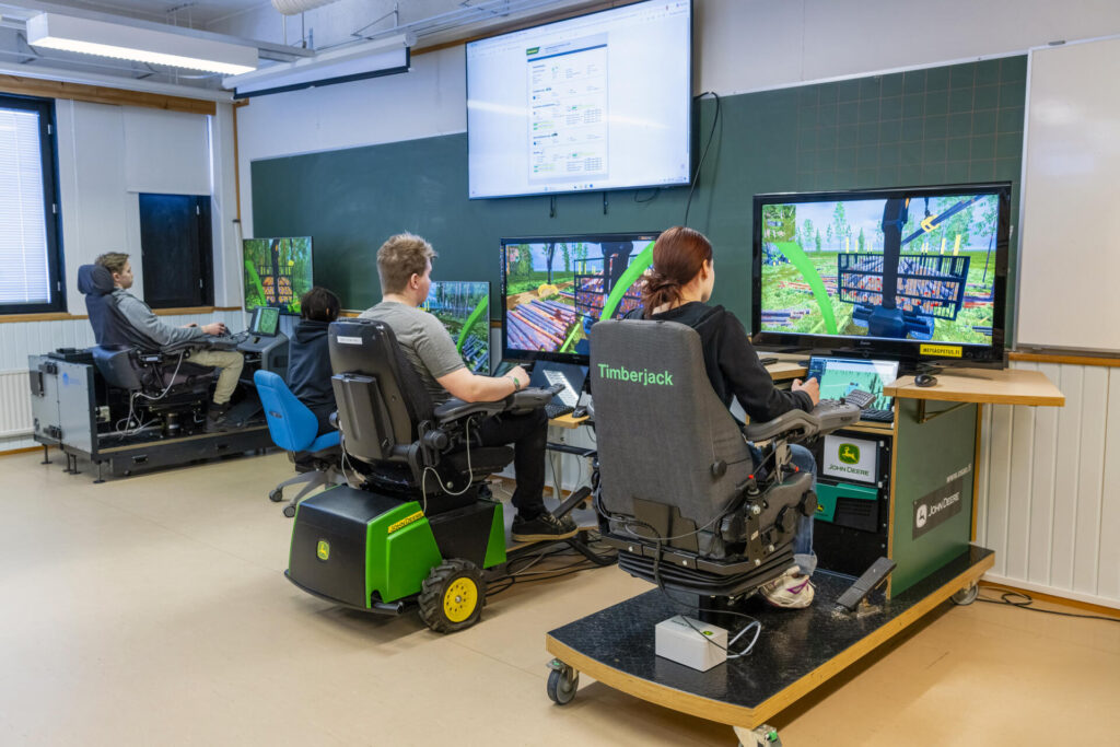 Opiskelijoita istumassa simulaattoreissa luokkahuoneessa. Näytöillä näkyy metsäkoneen simulaatioharjoitus metsänäkymässä. Seinällä on suuri näyttö, jossa on simulaation käyttöliittymä.