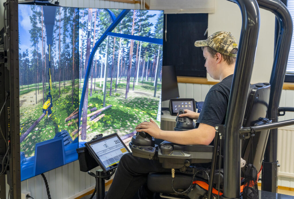 Henkilö istuu simulaattorissa ohjaamassa metsäkoneen toimintaa virtuaalisessa metsäympäristössä. Näytöllä näkyy metsänäkymä ja koneen käsittely puiden välissä.