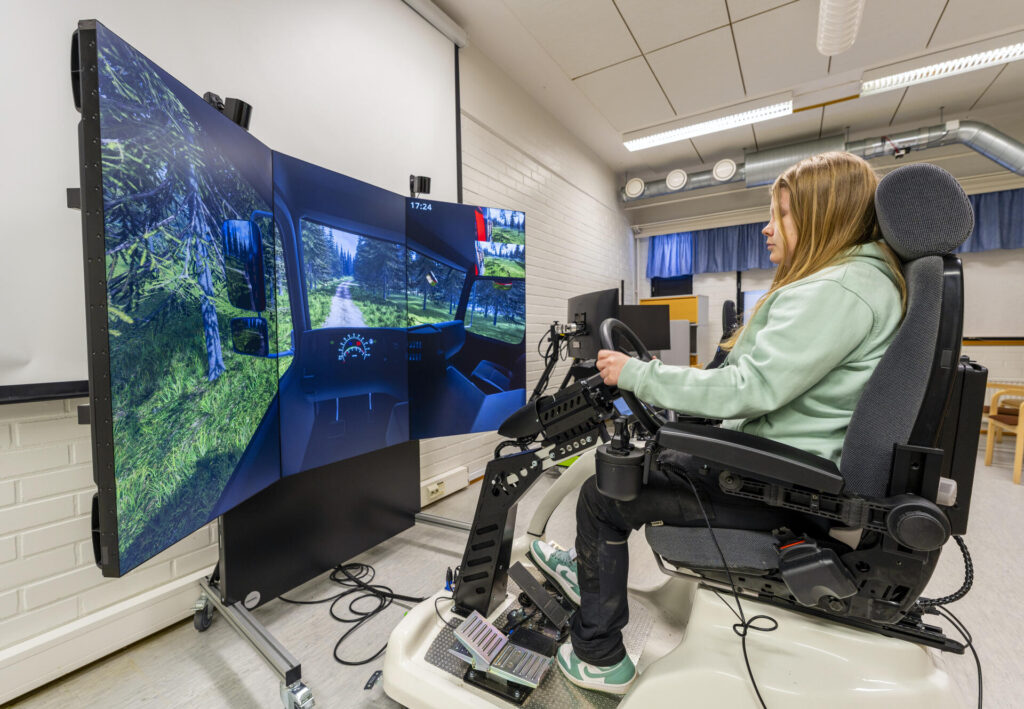 Opiskelija tekee harjoitusta metsäkonesimulaattorissa. Näytöllä näkyy metsämaisema ja ajoneuvon ohjaamo. Simulaattori sijaitsee sisätilassa, valkoisessa huoneessa, ja se koostuu useista näytöistä ja hallintalaitteista. Opiskelija istuu simulaattorin ohjaamossa ja ohjaa laitetta.