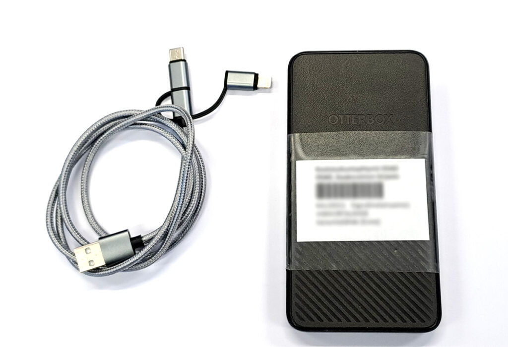 Kuvassa on musta ulkoinen akkupaketti, jossa on "OtterBox"-logo, ja sen vieressä on harmaa USB-kaapeli, jossa on useita liittimiä. Akkupaketin päällä on valkoinen tarra, jossa on sumennettu teksti.