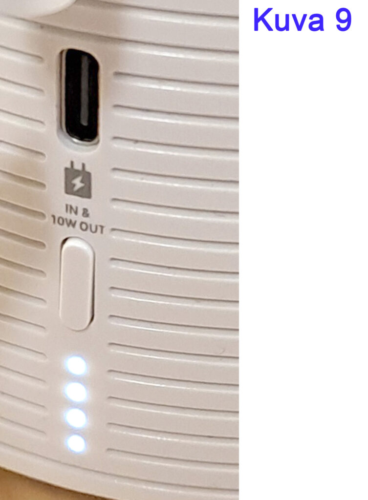 Lähikuva valkoisesta elektronisesta laitteesta, jossa on USB-portti ja kolme pientä valkoista LED-valoa, jotka osoittavat virran tai latauksen tason. Teksti "IN & 10W OUT" näkyy laitteen pinnalla.