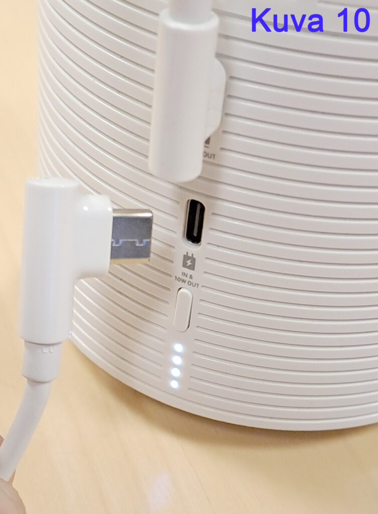 Lähikuva valkoisesta laitteesta, jossa on USB-C-portti ja siihen kytkettävä kaapeli. Laitteessa on myös pieniä LED-valoja, jotka osoittavat virran tilan. Kuvassa näkyy myös teksti 'Kuva 10' oikeassa yläkulmassa.