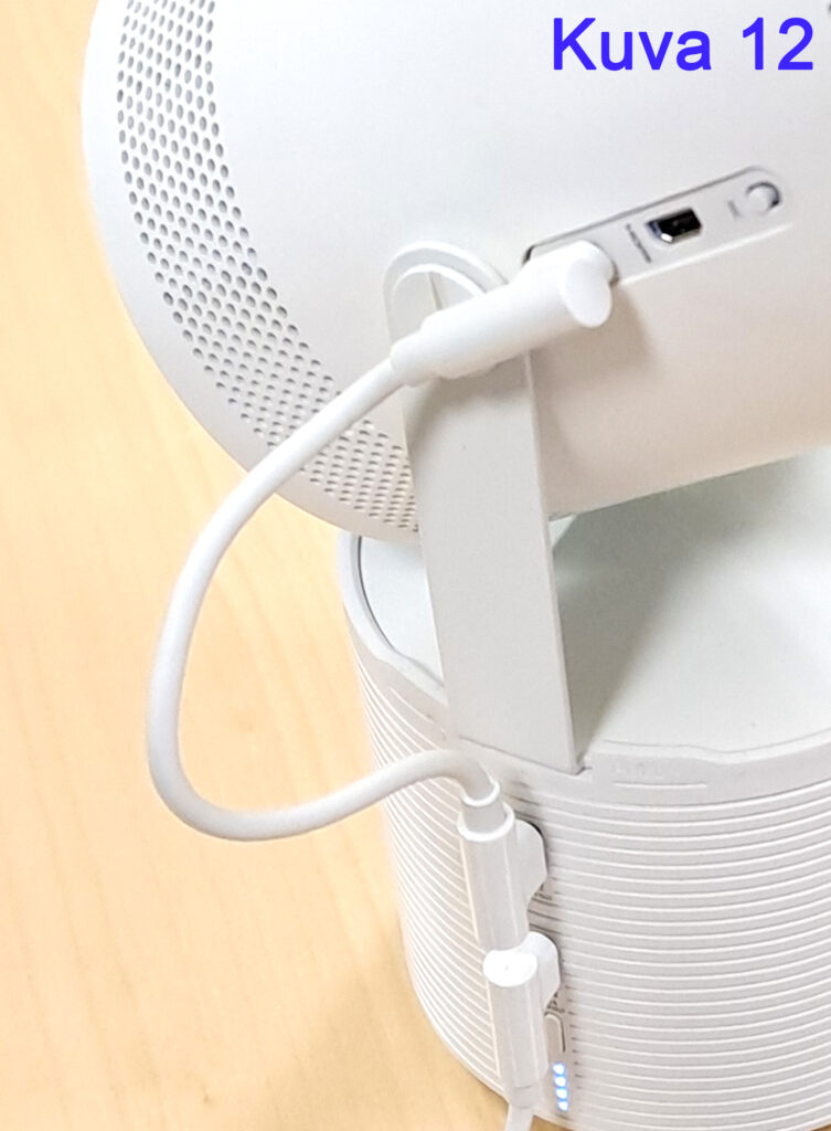 Lähikuva valkoisesta elektronisesta laitteesta, jossa on pyöreä kaiutin ja siihen liitetty valkoinen johto. "Kuva 12" on kirjoitettu sinisellä tekstillä kuvan oikeassa yläkulmassa.