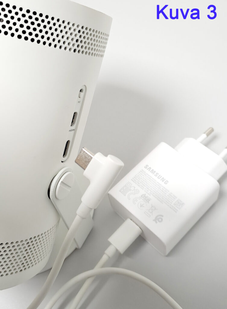 Lähikuva valkoisesta laitteesta, jossa on useita liitäntöjä kuten HDMI ja USB, sekä valkoinen Samsung-laturi, joka on liitetty laitteeseen.