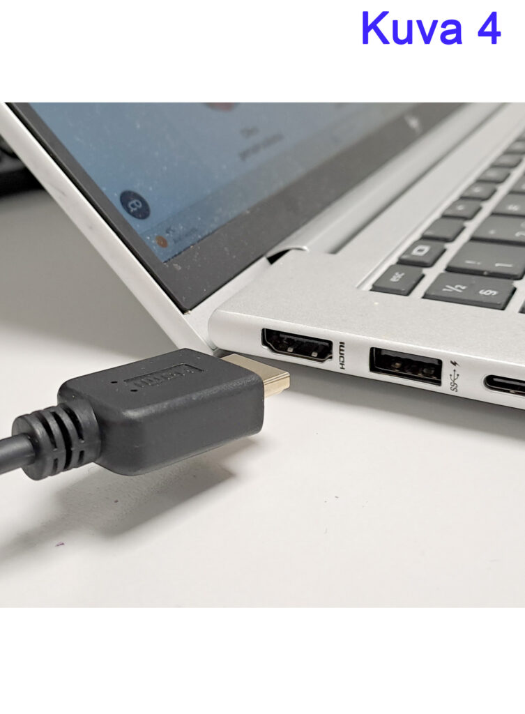 Kannettava tietokone, jossa näkyy HDMI-, USB- ja USB-C-portit, ja HDMI-kaapeli on kytkettynä lähes kiinni. Taustalla näkyy osittain kannettavan tietokoneen näyttö ja näppäimistö.