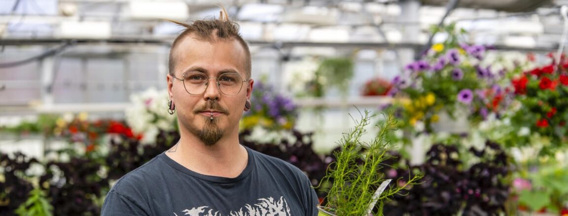 Henkilö seisoo kasvihuoneessa, pitäen vihreää kukkaruukkua kädessään. Hänen ympärillään on erilaisia kukkia ja kasveja. Henkilöllä on tatuointeja käsivarsissaan ja hänellä on silmälasit.