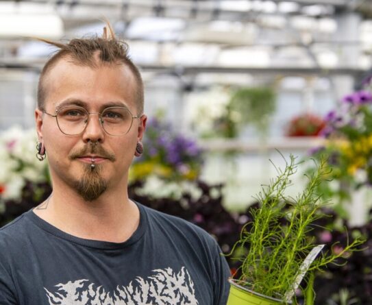 Henkilö seisoo kasvihuoneessa, pitäen vihreää kukkaruukkua kädessään. Hänen ympärillään on erilaisia kukkia ja kasveja. Henkilöllä on tatuointeja käsivarsissaan ja hänellä on silmälasit.