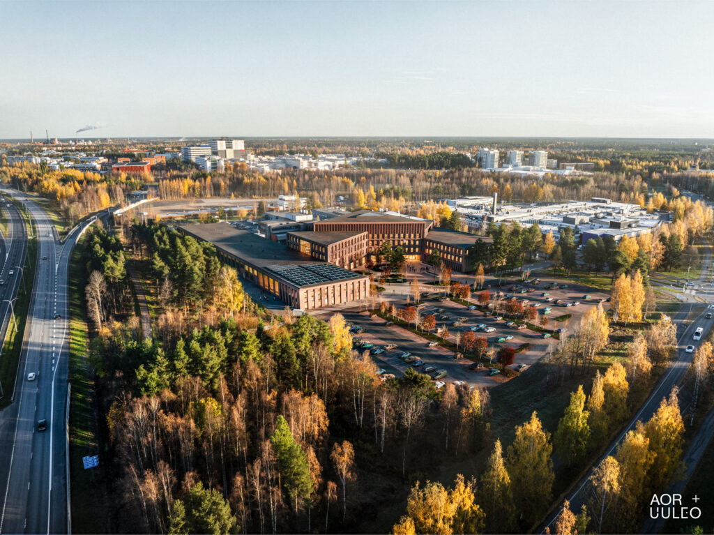 Ilmakuva modernista rakennuskompleksista syksyisessä maisemassa. Rakennukset ovat tiiliverhoiltuja ja sijaitsevat metsäisellä alueella, jota ympäröivät tiet ja parkkipaikat. Taustalla näkyy kaupunkirakennuksia ja syksyn värikkäitä puita.