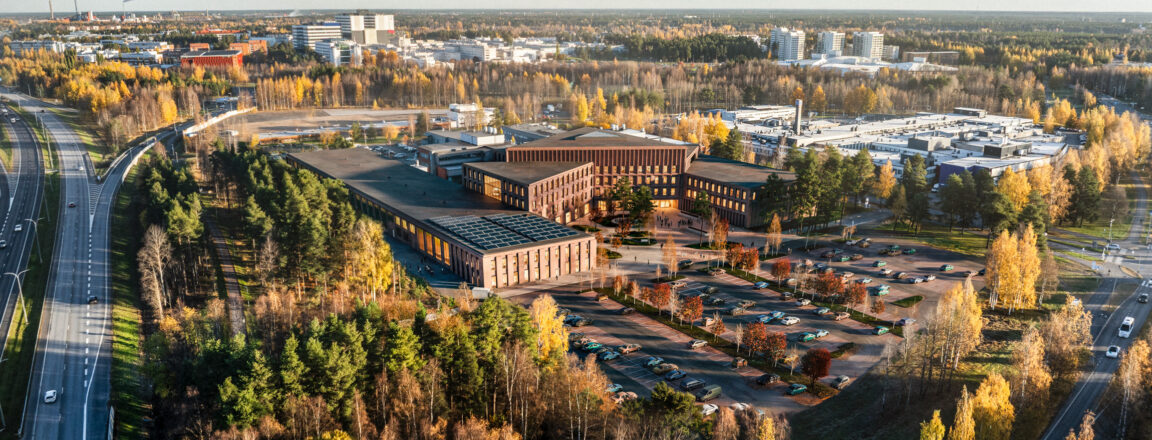Ilmakuva modernista rakennuskompleksista syksyisessä maisemassa. Rakennukset ovat tiiliverhoiltuja ja sijaitsevat metsäisellä alueella, jota ympäröivät tiet ja parkkipaikat. Taustalla näkyy kaupunkirakennuksia ja syksyn värikkäitä puita.