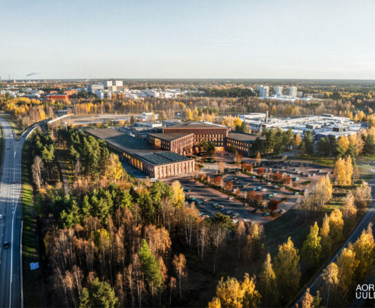 Ilmakuva modernista rakennuskompleksista syksyisessä maisemassa. Rakennukset ovat tiiliverhoiltuja ja sijaitsevat metsäisellä alueella, jota ympäröivät tiet ja parkkipaikat. Taustalla näkyy kaupunkirakennuksia ja syksyn värikkäitä puita.
