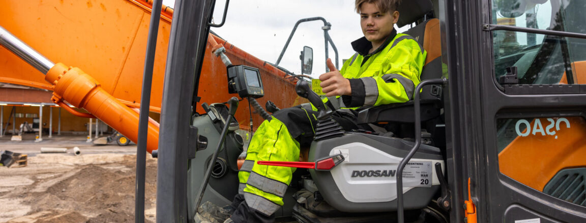 Nuori mies istuu kaivinkoneen ohjaamossa, pukeutuneena kirkkaan keltaiseen työtakkiin ja mustiin housuihin. Taustalla näkyy kaivinkoneen suuri oranssi puomi ja rakennustyömaa.