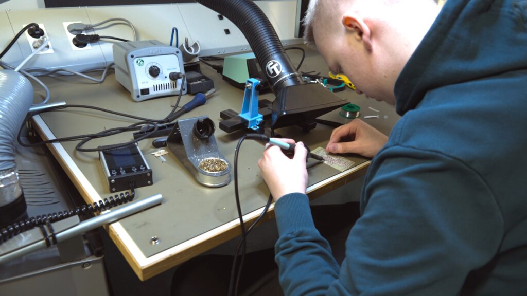 Henkilö työskentelee elektroniikkatyöpisteessä juottaen komponentteja piirikortille.