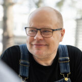 Mies, jolla on silmälasit ja musta paita, katsoo hymyillen sivulle. Hänellä on yllään siniset farkkuhaalarit.