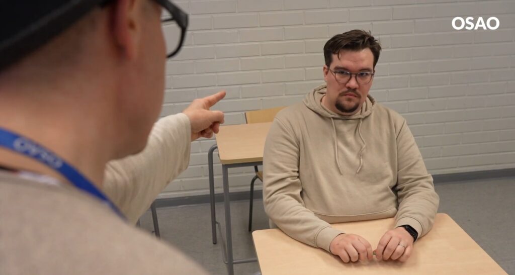 Kaksi henkilöä luokkahuoneessa. Toinen henkilö osoittaa sormellaan istuvaa henkilöä, jolla on vaalea huppari ja silmälasit.