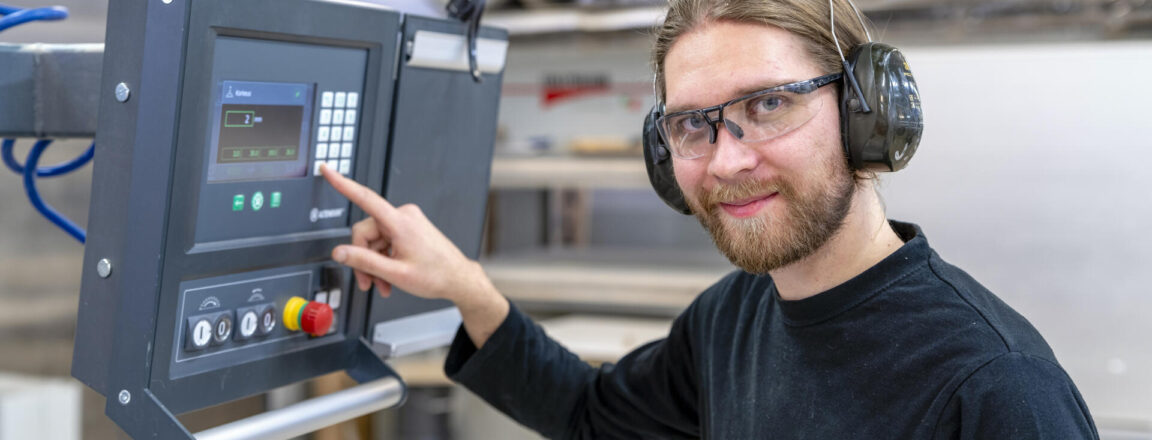 Mies seisoo teollisuuslaitteiston edessä, käyttäen kuulonsuojaimia ja suojalaseja. Hän osoittaa sormellaan ohjauspaneelin näyttöä.