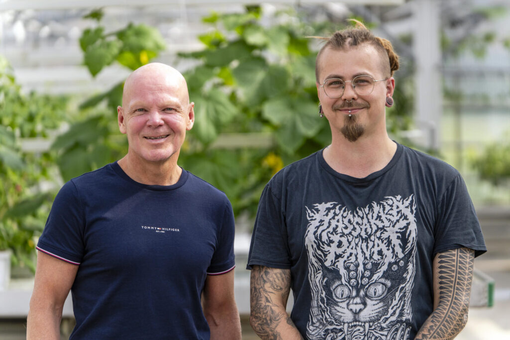 Kaksi miestä seisoo vierekkäin kasvihuoneessa kasvien ympäröimänä ja katsoo hymyillen kameraa kohti.