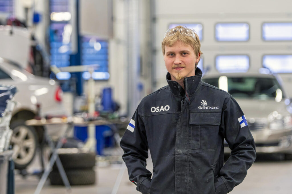 Nuori mies katsoo hymyillen kameraan, päällään työasu, jossa on OSAOn ja SkillsFinlandin logot ja hihoissa Suomen liput. Hän seisoo työsalissa, jossa on ajoneuvoja ja työkaluja taustalla.