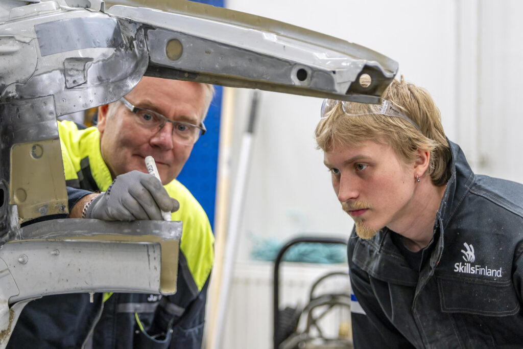 Kaksi miestä työskentelee yhdessä auton metallirakenteen kimpussa. Vasemmalla oleva mies pitelee kynää ja näyttää hymyilevän. Oikealla oleva mies katsoo keskittyneesti rakenteeseen. Heillä on työtakit päällä, ja taustalla näkyy työkaluja.