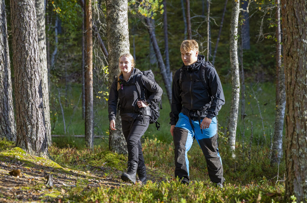Kaksi henkilöä kävelee metsässä selkäreput selässään, pukeutuneina ulkoiluvaatteisiin. He ovat keskellä vihreää metsää, ja auringonvalo siivilöityy puiden läpi.
