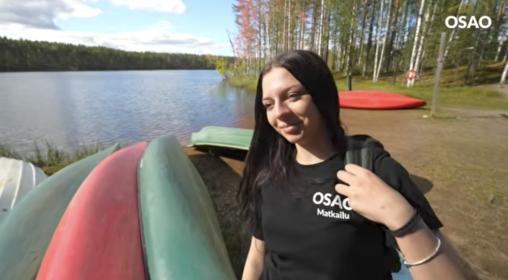 Nuori nainen seisoo järven rannalla, jossa on kumottuja kanootteja. Hänellä on musta OSAO-paita päällään ja reppu selässään. Taustalla näkyy vettä ja puita, joissa on syksyn väriset lehdet.