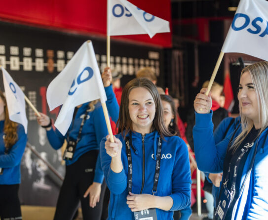 Ihmisiä sinisissä huppareissa kantamassa OSAO-lippuja sisätiloissa, taustalla seinä, jossa punaista ja mustaa väriä.