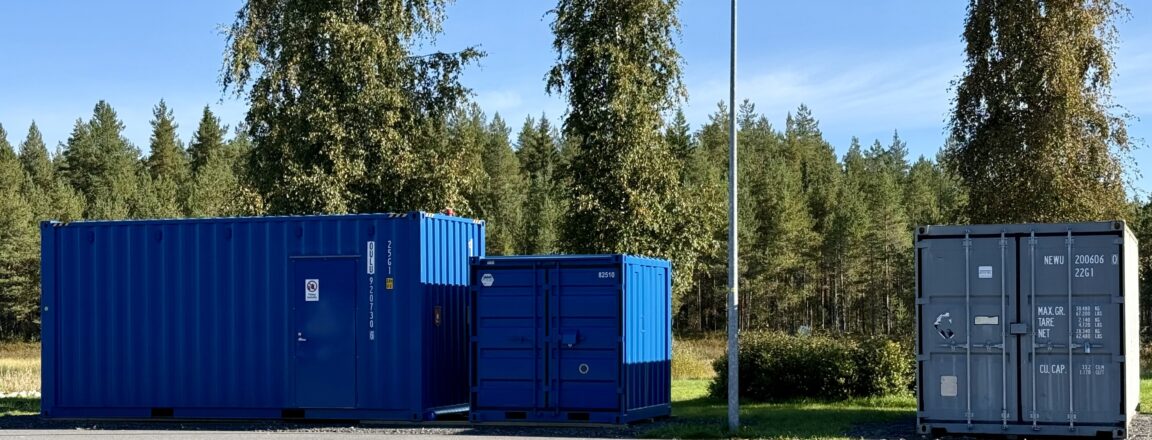 Kolme erikokoista konttia pihamaalla, jossa taustalla puita, pensaita ja kauempana metsää sekä sininen taivas. Kaksi konteista on sinisiä ja kolmas harmaa. Konttien välissä on katulamppu.