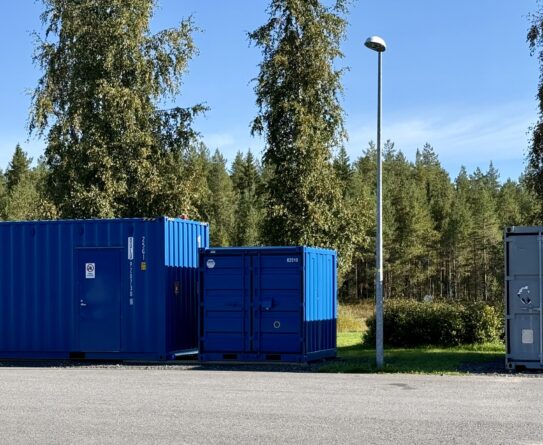 Kolme erikokoista konttia pihamaalla, jossa taustalla puita, pensaita ja kauempana metsää sekä sininen taivas. Kaksi konteista on sinisiä ja kolmas harmaa. Konttien välissä on katulamppu.