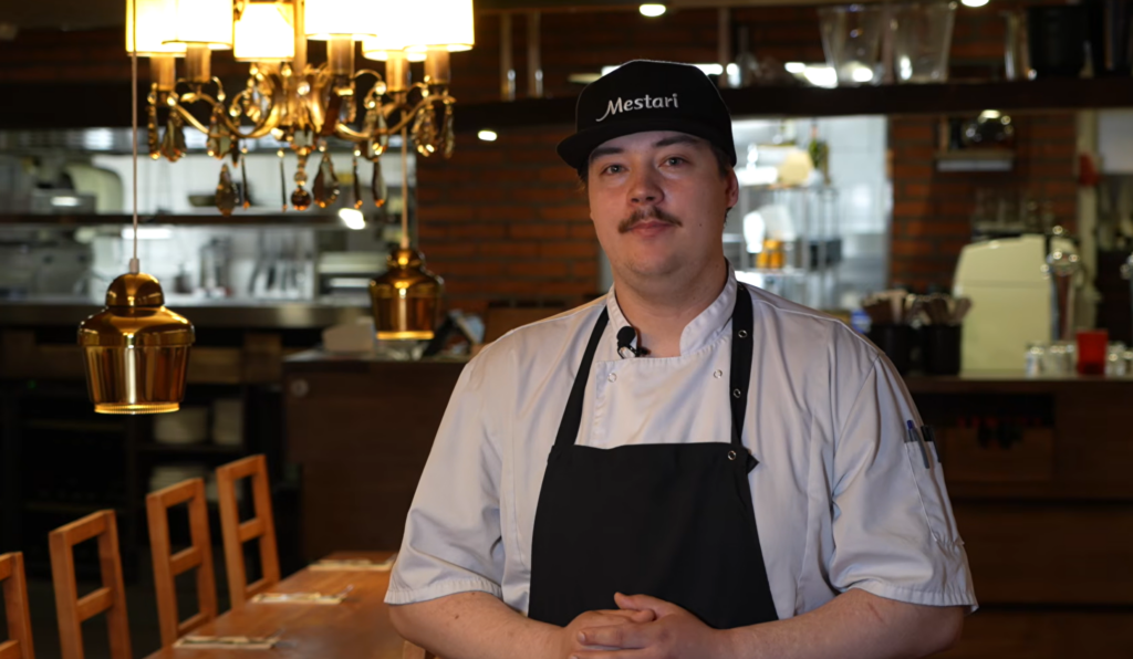 Kokki seisoo ravintolassa pukeutuneena kokin asuun ja lippalakkiin, jossa lukee "Mestari". Taustalla näkyy ravintolan sisustus, jossa on puupöytiä ja kattovalaisimia.